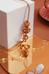 A gold chain with ornate details and small pearls is elegantly draped over a white geometric block. The jewelry reflects off a shiny, warm-toned surface and its intricate design is complemented by the soft lighting. In the background, there's a hint of a patterned, reddish-orange plate.
