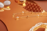 A close-up of elegant jewelry pieces, including a delicate gold chain with small pearls and a pair of ornate cufflinks set on a reflective gold surface. The background features intricate patterns that enhance the luxurious feel.