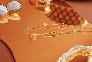 A close-up of elegant jewelry pieces, including a delicate gold chain with small pearls and a pair of ornate cufflinks set on a reflective gold surface. The background features intricate patterns that enhance the luxurious feel.