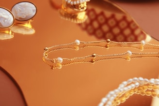 A close-up of elegant jewelry pieces, including a delicate gold chain with small pearls and a pair of ornate cufflinks set on a reflective gold surface. The background features intricate patterns that enhance the luxurious feel.