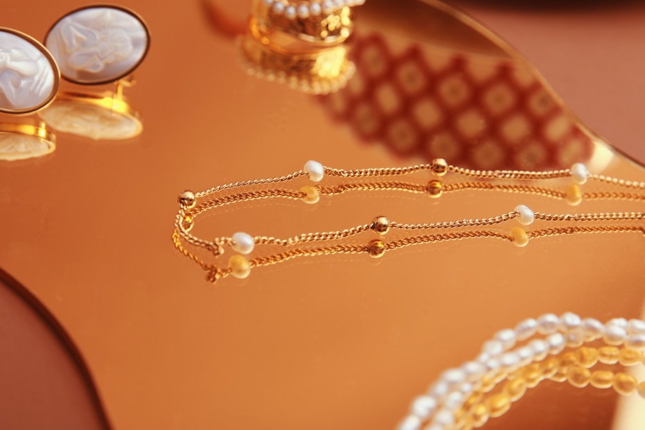 A close-up of elegant jewelry pieces, including a delicate gold chain with small pearls and a pair of ornate cufflinks set on a reflective gold surface. The background features intricate patterns that enhance the luxurious feel.