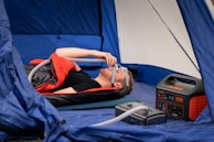 A person is lying inside a tent on a mattress, using a CPAP machine connected to a power station. They are covered with a sleeping bag, and there is camping gear neatly arranged around them.
