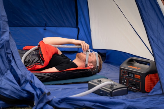 A person is lying inside a tent on a mattress, using a CPAP machine connected to a power station. They are covered with a sleeping bag, and there is camping gear neatly arranged around them.