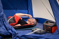 A person is lying inside a tent on a mattress, using a CPAP machine connected to a power station. They are covered with a sleeping bag, and there is camping gear neatly arranged around them.