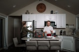 A modern kitchen interior features a man cooking on a countertop while a woman reaches into an open refrigerator. White cabinets and a large wall clock add to the contemporary feel. A portable power station is prominently placed on the counter next to a red pot.