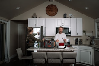 A modern kitchen interior features a man cooking on a countertop while a woman reaches into an open refrigerator. White cabinets and a large wall clock add to the contemporary feel. A portable power station is prominently placed on the counter next to a red pot.