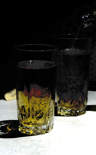 Two tall, clear glasses filled with a bubbly, golden liquid are placed against a dark background. One glass is being filled from a bottle, with the liquid visibly pouring into it and creating fizz. The lighting creates a strong contrast, highlighting the glasses and casting intricate shadows on the surface.