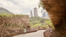 A person with a green backpack walks along a pathway flanked by lush greenery and arched wooden structures. The background features a city skyline, with several modern high-rise buildings under construction. The sky is overcast, adding a calm and serene atmosphere to the scene.