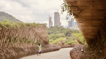 A person with a green backpack walks along a pathway flanked by lush greenery and arched wooden structures. The background features a city skyline, with several modern high-rise buildings under construction. The sky is overcast, adding a calm and serene atmosphere to the scene.