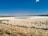 A vast salt flat with a small pool of water in the center under a clear blue sky. The terrain is rough and dry, with signs of salt crystallization. A sign in Spanish saying 'No Pasar' is visible in the foreground, indicating restricted access.