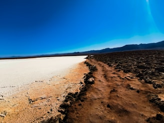 A rugged landscape with a clear division between a white salt flat and a brown, rocky surface, leading towards distant mountains under a bright blue sky.