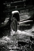 Jeep crossing a shallow river with splashing water under a bright blue sky.