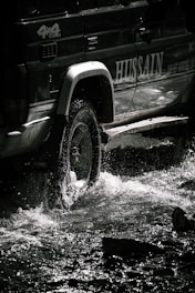 A rugged off-road vehicle is traversing through shallow water, causing splashes around its wheels. The vehicle bears noticeable branding on its side, with sunlight casting dramatic shadows.