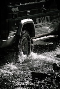 A rugged off-road vehicle is traversing through shallow water, causing splashes around its wheels. The vehicle bears noticeable branding on its side, with sunlight casting dramatic shadows.