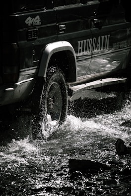 Jeep crossing a shallow river with splashing water under a bright blue sky.