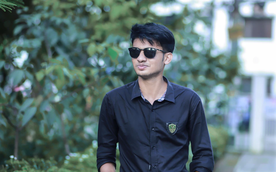 Young man stands on sidewalk wearing sunglasses and black shirt with green emblem