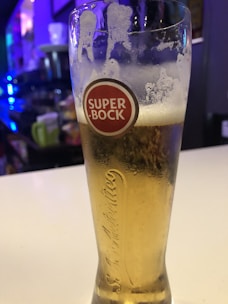 A tall glass filled with light beer sits on a counter. The glass is branded with the label 'Super Bock' in a red circle near the top. The beer appears cold with condensation on the outside of the glass, and remnants of foam cling to the inside of the rim. The background is slightly blurred, showing bar elements with hints of blue and purple ambient lighting.