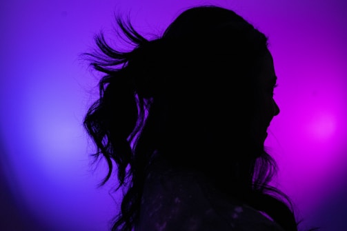 A silhouette of a person against a vibrant background with shades of purple and blue. The person's hair is styled in wavy locks tied back, and their facial features are darkened due to the backlighting.