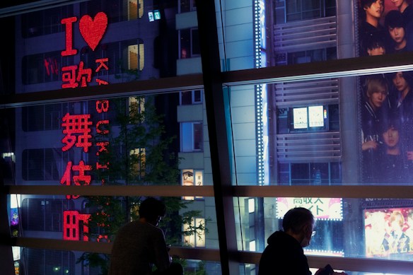 A nighttime urban scene with a brightly lit building showcasing vibrant signage. The neon text on the building includes a large illuminated heart symbol, surrounded by Japanese characters. Silhouettes of two people sitting by a large window frame the foreground, with one person noticeably using a phone. In the background, a large poster displays a group of individuals, partially visible.