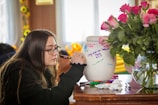 A person is writing on a white urn with colorful markers, which has various messages and decorations. A vase with pink roses is nearby on the wooden table. The setting suggests an intimate, reflective atmosphere.