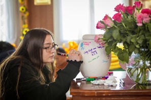 A person is writing on a white urn with colorful markers, which has various messages and decorations. A vase with pink roses is nearby on the wooden table. The setting suggests an intimate, reflective atmosphere.