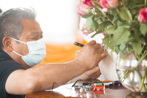 A person wearing a face mask is writing on a white surface with a pen. Several markers are placed on the table nearby. A vase with pink and white roses is prominently positioned to the right of the person.