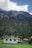 A small house with a flat roof, adorned with prayer flags, sits in a lush green valley. Behind it, a dense forest of tall pine trees stretches up the slope of a towering mountain range with rocky, jagged peaks under a partly cloudy blue sky.
