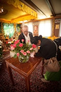 An indoor setting with two people by a wooden table decorated with a vase of pink roses. The room is ornately furnished with religious imagery, including framed pictures on the wall. A woman leans over the table, bowing her head, while the man stands beside her observing. The ceiling and walls have golden yellow drapery and decorations.