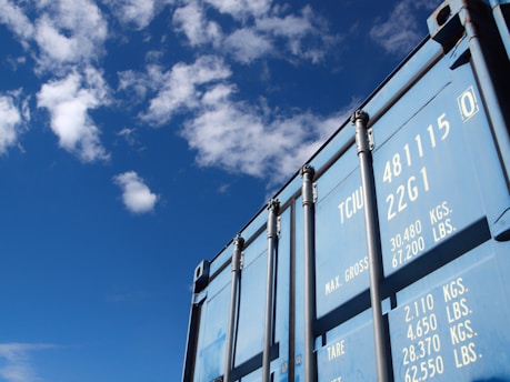 A blue shipping container is positioned against a bright blue sky with scattered clouds. The container has visible numbers and text printed in white on its surface, including weight specifications.