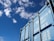 A blue shipping container is positioned against a bright blue sky with scattered clouds. The container has visible numbers and text printed in white on its surface, including weight specifications.