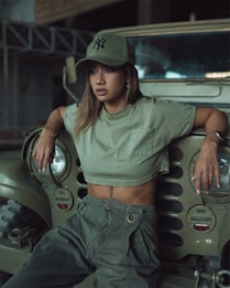 A person wearing an olive green cap and matching clothing is leaning against a vintage army-style jeep. The image has an urban and rugged aesthetic, with industrial elements in the background.