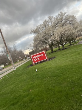A red KeyBank sign stands prominently on well-maintained green grass. In the background, there are several blossoming trees and a road with a few vehicles. The sky is overcast with dark, heavy clouds, creating a moody atmosphere.