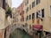 A narrow canal flows between two rows of historic buildings with shuttered windows. A small arched stone bridge crosses over the canal. Flags and banners hang from the buildings, and a restaurant canopy is visible with the name 'Ristorante Ivo'. Plants decorate some windowsills, adding a touch of greenery.