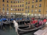 A row of gondolas docked in a canal by a hotel with a yellow facade. The windows have red awnings, and the building is illuminated, reflecting a warm glow onto the water. The gondolas have ornate decorations and various colored covers.