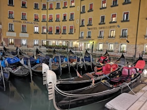 A row of gondolas docked in a canal by a hotel with a yellow facade. The windows have red awnings, and the building is illuminated, reflecting a warm glow onto the water. The gondolas have ornate decorations and various colored covers.