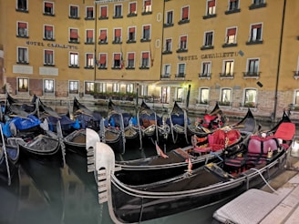 A row of gondolas docked in a canal by a hotel with a yellow facade. The windows have red awnings, and the building is illuminated, reflecting a warm glow onto the water. The gondolas have ornate decorations and various colored covers.
