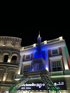 A nighttime scene featuring a building with architectural elements reminiscent of classical Roman and Parisian styles. The structure includes a model of the Eiffel Tower, illuminated in blue, and an adjoining facade resembling the Colosseum, lit with warm lights. The sign below reads 'Luxury Garments' with additional text in a different script.