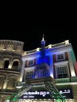 A nighttime scene featuring a building with architectural elements reminiscent of classical Roman and Parisian styles. The structure includes a model of the Eiffel Tower, illuminated in blue, and an adjoining facade resembling the Colosseum, lit with warm lights. The sign below reads 'Luxury Garments' with additional text in a different script.