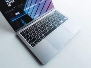 A laptop with a silver body and a black keyboard is open on a light surface. The screen displays a video editing program, showing timelines and clips. Natural light casts shadows and reflections on the screen.