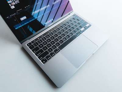 A laptop with a silver body and a black keyboard is open on a light surface. The screen displays a video editing program, showing timelines and clips. Natural light casts shadows and reflections on the screen.