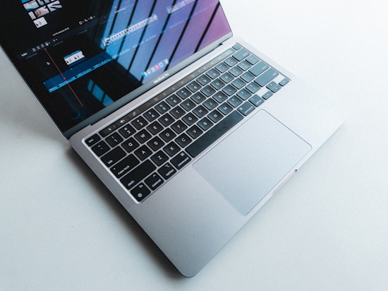 A laptop with a silver body and a black keyboard is open on a light surface. The screen displays a video editing program, showing timelines and clips. Natural light casts shadows and reflections on the screen.