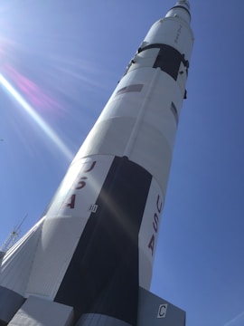 A large rocket standing upright against a clear blue sky. The rocket is painted white with black and red accents, and the word 'USA' is prominently displayed along with a U.S. flag. Sunlight creates a lens flare across the image.