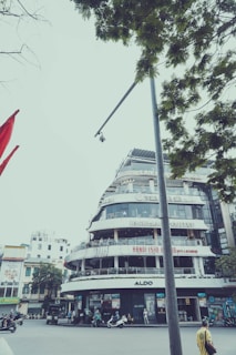 A multi-story building featuring several shops and cafes, prominently displaying signs such as 'Highlands Coffee' and 'Aldo'. The architecture is modern with rounded corners and balconies. Below, there are people and motorcycles, indicating a busy urban environment. Trees with green leaves partially frame the top of the scene, and a camera is mounted on a tall pole in the foreground.