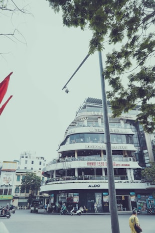 A multi-story building featuring several shops and cafes, prominently displaying signs such as 'Highlands Coffee' and 'Aldo'. The architecture is modern with rounded corners and balconies. Below, there are people and motorcycles, indicating a busy urban environment. Trees with green leaves partially frame the top of the scene, and a camera is mounted on a tall pole in the foreground.