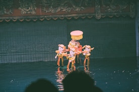 Wooden puppets dressed in traditional attire are performing on water. The puppets appear to be carrying a decorated umbrella-like object, and the background is an intricately designed stage.