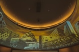 A curved panoramic display features a series of modern architectural buildings, illuminated by a warm yellow-orange light. The structures exhibit diverse architectural styles, with geometric designs and an array of windows. The setting appears to be an interior space designed for showcasing architectural imagery.