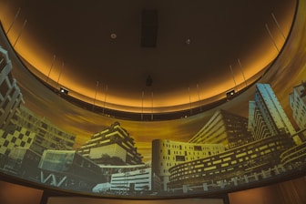 A curved panoramic display features a series of modern architectural buildings, illuminated by a warm yellow-orange light. The structures exhibit diverse architectural styles, with geometric designs and an array of windows. The setting appears to be an interior space designed for showcasing architectural imagery.