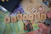 Scrabble tiles spelling out 'rebates' are arranged on top of various Canadian banknotes including denominations like 20 and 100. A part of a passport is visible in the corner.