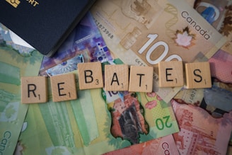 Scrabble tiles spelling out 'rebates' are arranged on top of various Canadian banknotes including denominations like 20 and 100. A part of a passport is visible in the corner.