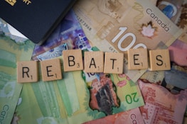 Scrabble tiles spelling out 'rebates' are arranged on top of various Canadian banknotes including denominations like 20 and 100. A part of a passport is visible in the corner.